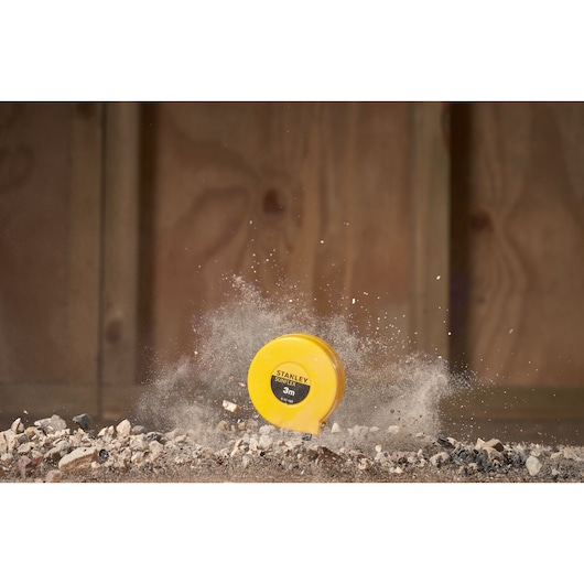 STANLEY 0-32-189 tape measure in yellow casing on rubble, impact dust in background, photography.