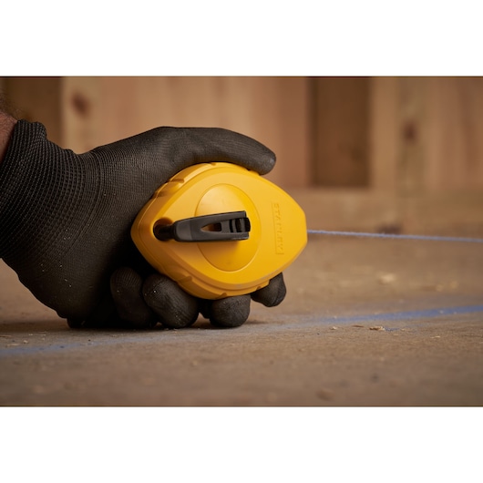 Photo of STANLEY 0-47-440 chalk line reel in use, yellow casing, black gloved hand marking a surface.
