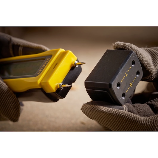 STANLEY 0-77-030 digital moisture meter close-up with probe points and calibration block held by gloved hands.