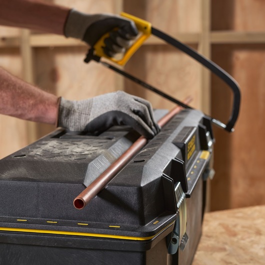 STANLEY 1-93-935 toolbox used as work surface, gloved hands cutting copper pipe with hacksaw, front angle