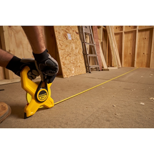Photo of STANLEY 2-34-792 tape measure with 30m yellow blade in use on construction floor, gloved hand holding tool.
