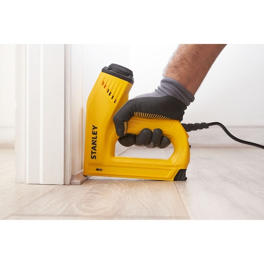 Photo of STANLEY 6-TRE550 electric stapler in use near baseboard, yellow body, side angle view.