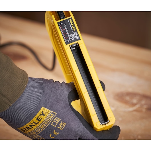 STANLEY 6-TRE550 electric stapler close-up held in gloved hand, yellow body, wood surface underneath.