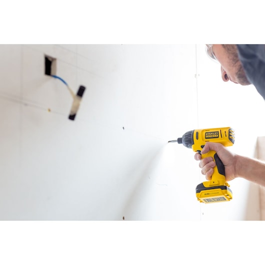 Stanley FMC041S2 cordless drill in use, yellow color, shown drilling into white drywall.