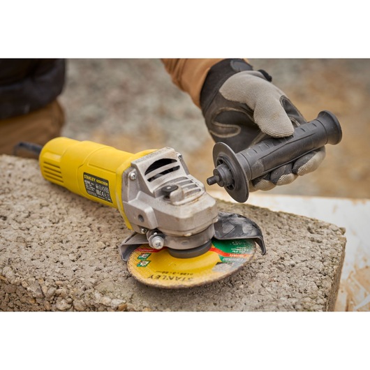 STANLEY FMEG615 angle grinder with yellow body, worker attaching side handle, outdoor concrete surface.
