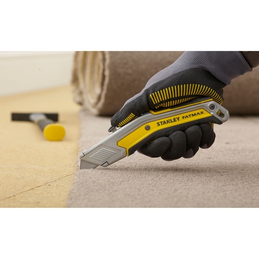 STANLEY FMHT0-10288 utility knife cutting carpet, ergonomic yellow and silver handle, gloved hand holding tool.