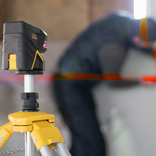 STANLEY FMHT1-77414 laser level on tripod, emitting red lines, indoor workspace, angled side view.
