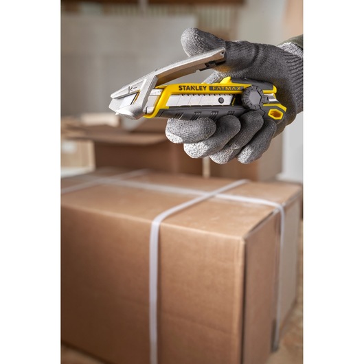 STANLEY FMHT10592-0 utility knife held in gloved hand, yellow and black, with cardboard boxes in background.