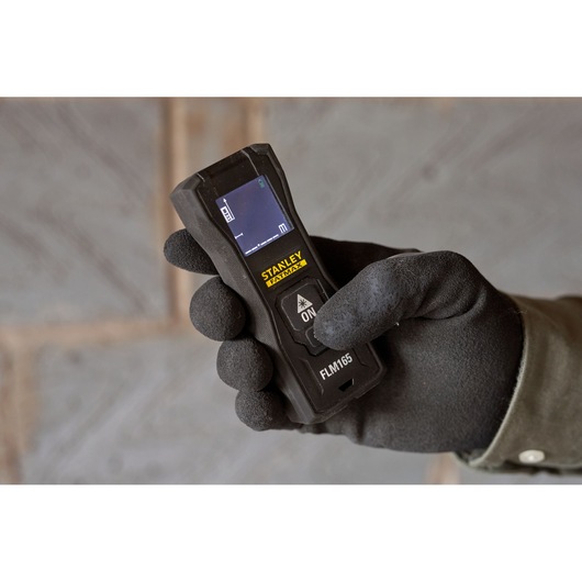 Photo of STANLEY FMHT77165-0 laser distance measurer held in a gloved hand, screen and buttons visible.