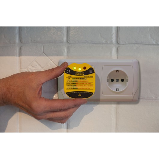 STANLEY FMHT82569-6 socket tester held near white outlet, yellow casing, lights and indicator labels visible.