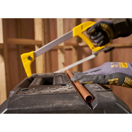 STANLEY FMST1-71219 photography of a gloved hand using a hacksaw to cut a copper pipe on a black workbench.
