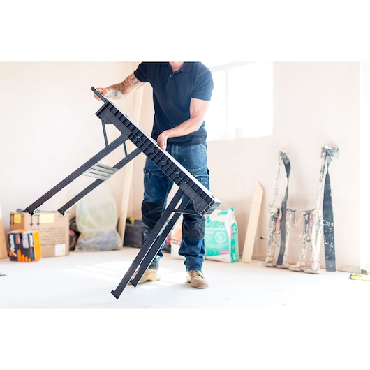 Person setting up a STANLEY FMST1-75672 folding sawhorse in a bright workspace.