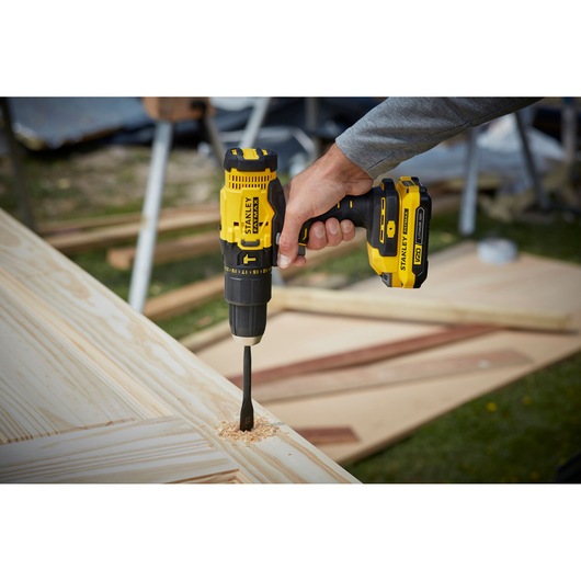 Photo of STANLEY SFMCD711C2S cordless drill in use, drilling into light wood, outdoor worksite background.