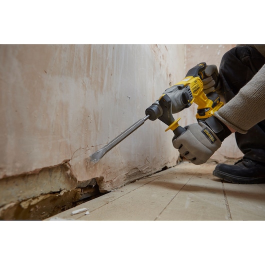 STANLEY SFMCH900M22 rotary hammer in use chipping plaster from a wall near the floor, close-up action shot.