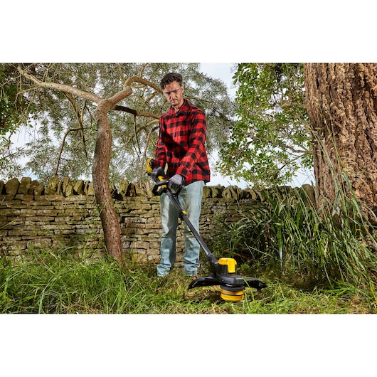 STANLEY SFMCSTB933B string trimmer in use outdoors, yellow and black, grass cutting near stone wall.