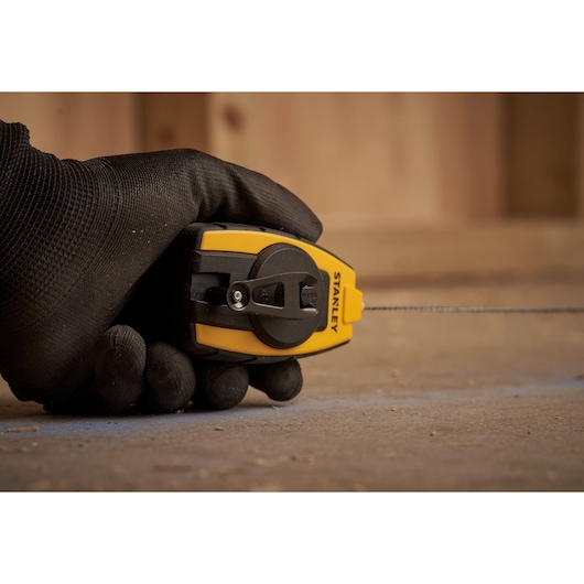 Photo of STANLEY STHT0-47147 chalk line reel held in a gloved hand, yellow and black, marking a surface.