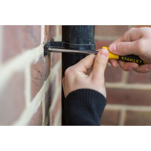 Hand uses STANLEY screwdriver to tighten screw on pipe bracket attached to brick wall.