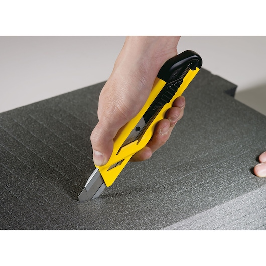 Photo of STANLEY STHT10265-0 utility knife cutting black foam sheet, close-up view of hand holding yellow knife.