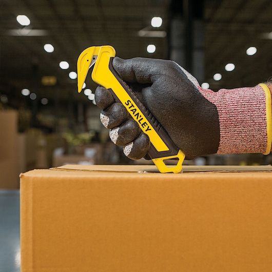 Stanley STHT10356 box cutter held with glove, cutting cardboard box in warehouse setting.