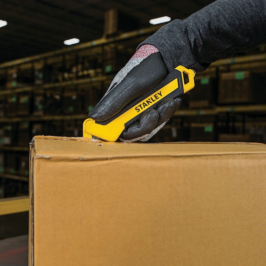 Photo of STANLEY STHT10356 box cutter in use, yellow and black, opening a cardboard box in warehouse setting.