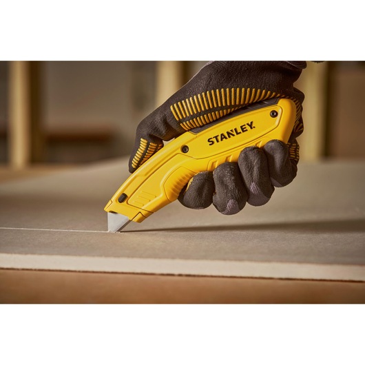 Photo of STANLEY STHT10430-0 utility knife cutting drywall, held by gloved hand.