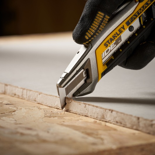 Photo of STANLEY STHT2-11818 utility knife cutting drywall, close-up on blade and hand in glove.