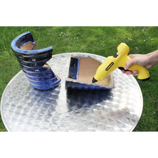 Photo of a STANLEY STHT6-70416 glue gun being used to repair a broken blue ceramic pot on a metal table outdoors.