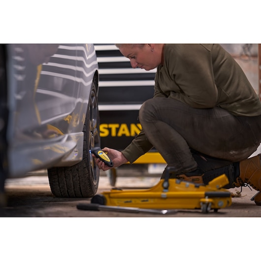 Person using STANLEY STHT80874-0 digital tire pressure gauge on car wheel, yellow tools and jack on ground.